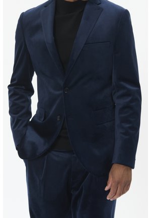 Mann trägt eine dunkelblaue Samtsakko und passende Hose mit einem schwarzen Hemd darunter, steht mit einer Hand in der Tasche.