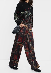 Maglione nero con ricami floreali; pantaloni a gamba larga con motivo e accenti rossi e dorati; abbinati a calzature nere e una borsa piccola.