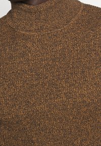 Gros plan sur un pull à col roulé tricoté marron et noir porté par une personne à la peau foncée.