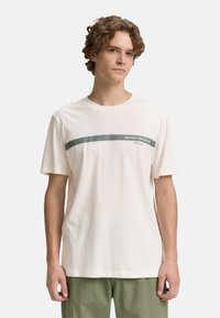 Young man with curly hair wears a light-colored t-shirt featuring a green stripe and text, standing against a plain white background.