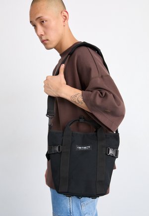 Ung person bærer en sort Carhartt skuldertaske, iført en brun oversized skjorte og lyseblå jeans, med en synlig tatovering på håndleddet.