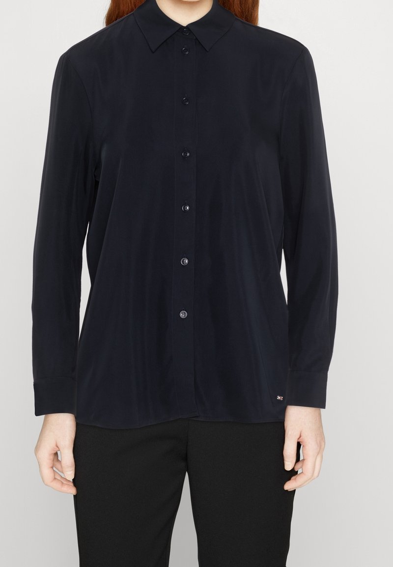 Femme portant une chemise noire à manches longues avec col et boutons et un pantalon noir, debout devant un fond uni.