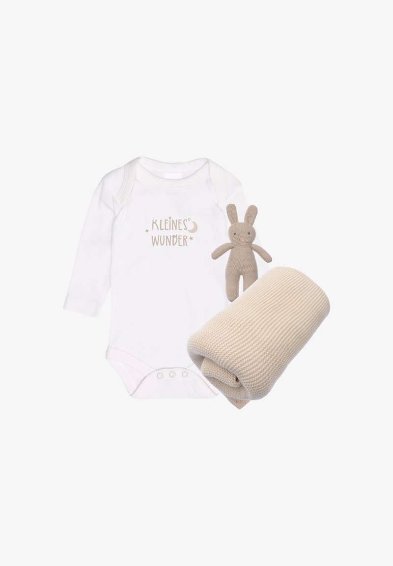 Weißer Langarm-Overall mit dem Text "KLEINES WUNDER", einem beigen Plüschhasen und einer gerollten, waffle-texturierten beigen Decke.