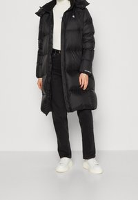 Černý péřový kabát s kapucí, dlouhý střih, prošívaný design a s logem, doplněný černými kalhotami a bílými teniskami.