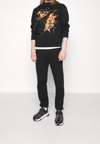 Schwarzer Hoodie mit lebhaftem Grafikdesign, kombiniert mit schwarzen Sweatpants und schwarzen Sportschuhen mit weißen Akzenten und Schnürsenkeln. Umhängetasche inklusive.