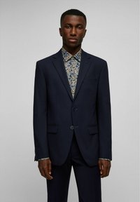 Marineblazer met twee knopen, schuin geplaatste klepzakken en blauwe accenten. Daaronder een gestreept overhemd met blauwe en gouden bloemenmotieven.