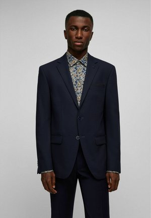 Marineblazer met twee knopen, schuin geplaatste klepzakken en blauwe accenten. Daaronder een gestreept overhemd met blauwe en gouden bloemenmotieven.
