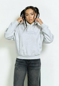 Gray hoodie with a front pocket and hood, featuring a decorative text in rhinestones. Styled with dark jeans, set against a neutral background.