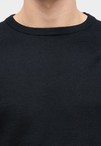 Gros plan sur le cou et le haut de la poitrine d’une personne vêtue d’un pull noir uni à col rond, sur un fond clair.