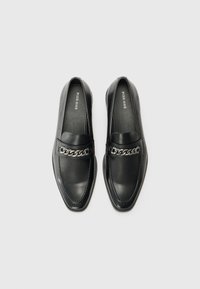 Mocassins en cuir noir avec un design plat et un bout pointu. Dotés d'une chaîne décorative en argent sur le devant, d'une texture lisse et de coutures discrètes.