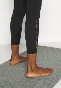 Leggings neri con un design aderente e tessuto testurizzato, caratterizzati da dettagli con ritagli nella parte inferiore della gamba. Indossati a piedi nudi su un tappetino grigio.