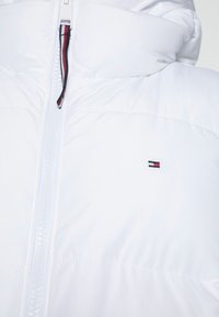 Chaqueta de plumas blanca con cremallera frontal y pequeño logo rojo, blanco y azul en el pecho, y tirador de la cremallera con detalle a rayas a juego.