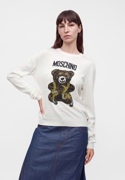 Femme aux cheveux bruns portant un pull blanc Moschino avec un motif d'ours en peluche et une jupe en jean bleu, debout avec une main sur la hanche.