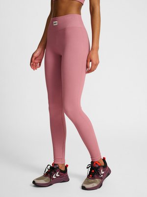 Person, die hoch taillierte, gerippte pinkfarbene Leggings und mehrfarbige Laufschuhe trägt, steht vor einem einfarbigen hellen Hintergrund.