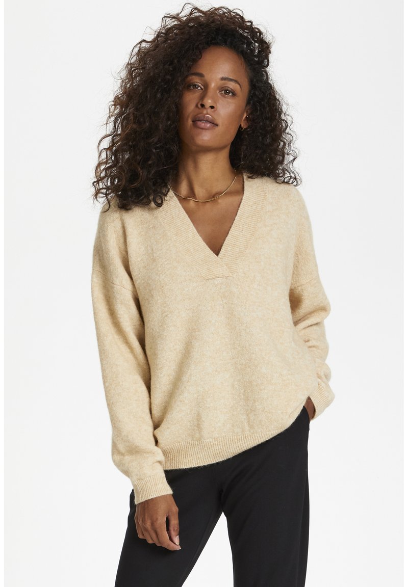 Pull en v beige surdimensionné en tissu doux et texturé. Il a des manches longues et des poignets et un ourlet côtelés. Porté avec un pantalon sombre.