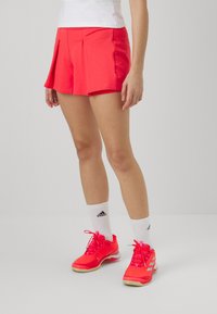 Pantalones cortos deportivos de color rosa brillante con pliegues, hechos de un tejido ligero. Se llevan con calcetas deportivas blancas y zapatillas rosas a juego.
