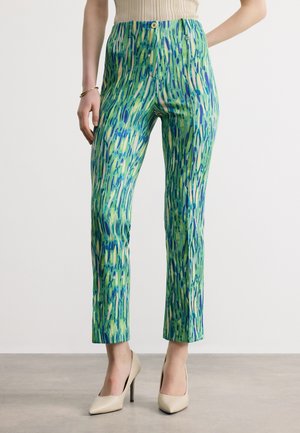 Frau trägt grüne und blaue abstrakte, figurbetonte Knöchellange Hose, kombiniert mit beigen High-Heels und einem beige ärmellosen Oberteil.