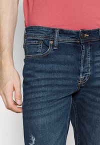 Jeans de mezclilla azul oscuro con un ligero desgaste, que presentan un diseño clásico de cinco bolsillos, costuras amarillas y un cierre de botón único en la cintura.