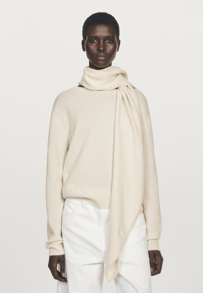 Model with short hair wears cream sweater, matching long scarf draped over shoulder, and white pants against plain light background.