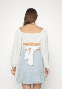Bílý texturovaný crop top s dlouhými puff rukávy a zadním šněrováním, spárovaný s světle modrou květinovou mini sukní s volánkovým lemem.