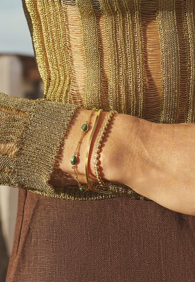 Mâneca tricotată din aur cu un material texturat maro dedesubt. Trei brățări: aurie cu pietre verzi, lanț de aur și una cu mărgele.
