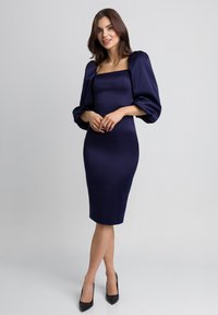 Niebieska satynowa sukienka z kwadratowym dekoltem i bufkami, dopasowana sylwetka, midi długość, zestawiona z czarnymi szpilkami w ostrym nosku.