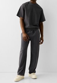 Šedé oversized tričko vyrobené z bavlněné směsi s krátkým rukávem, spárované s volnými šedými kalhotami. Oba kousky mají hladkou texturu a jednoduchý design.