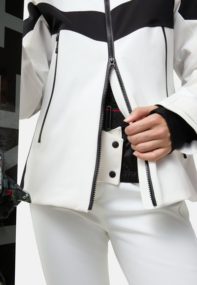 Hvid og sort zip-jakke med en struktureret overflade, der har kontrasterende farver og en frontlynlås, kombineret med tætsiddende hvide bukser.
