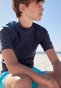 Tumesinist lühikeste varrukatega rüübikaitse, mis on valmistatud siledast ja elastsest materjalist, kombineeritud helesiniste ujumispükstega, kandmas noor inimene rannas.