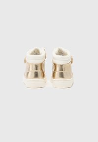 Baskets montantes avec un dos métallique doré brillant, un col rembourré blanc et une semelle texturée blanche. Sangle Velcro sur le côté.