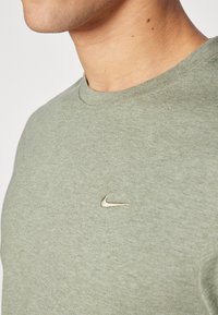 Olíva zöld, rövid ujjú póló, kerek nyakkivágással; a mellkason egy kis arany Nike logó található; puha anyagból készült.