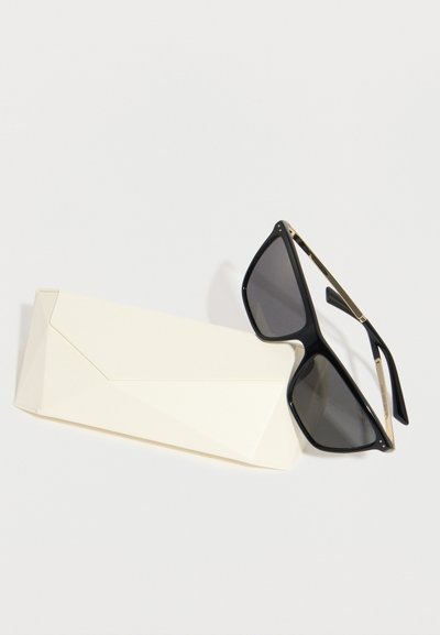 Μαύρα ορθογώνια γυαλιά ηλίου με χρυσές λεπτομέρειες που ακουμπούν σε μια κλειστή λευκή ορθογώνια θήκη γυαλιών σε απλή λευκή επιφάνεια.