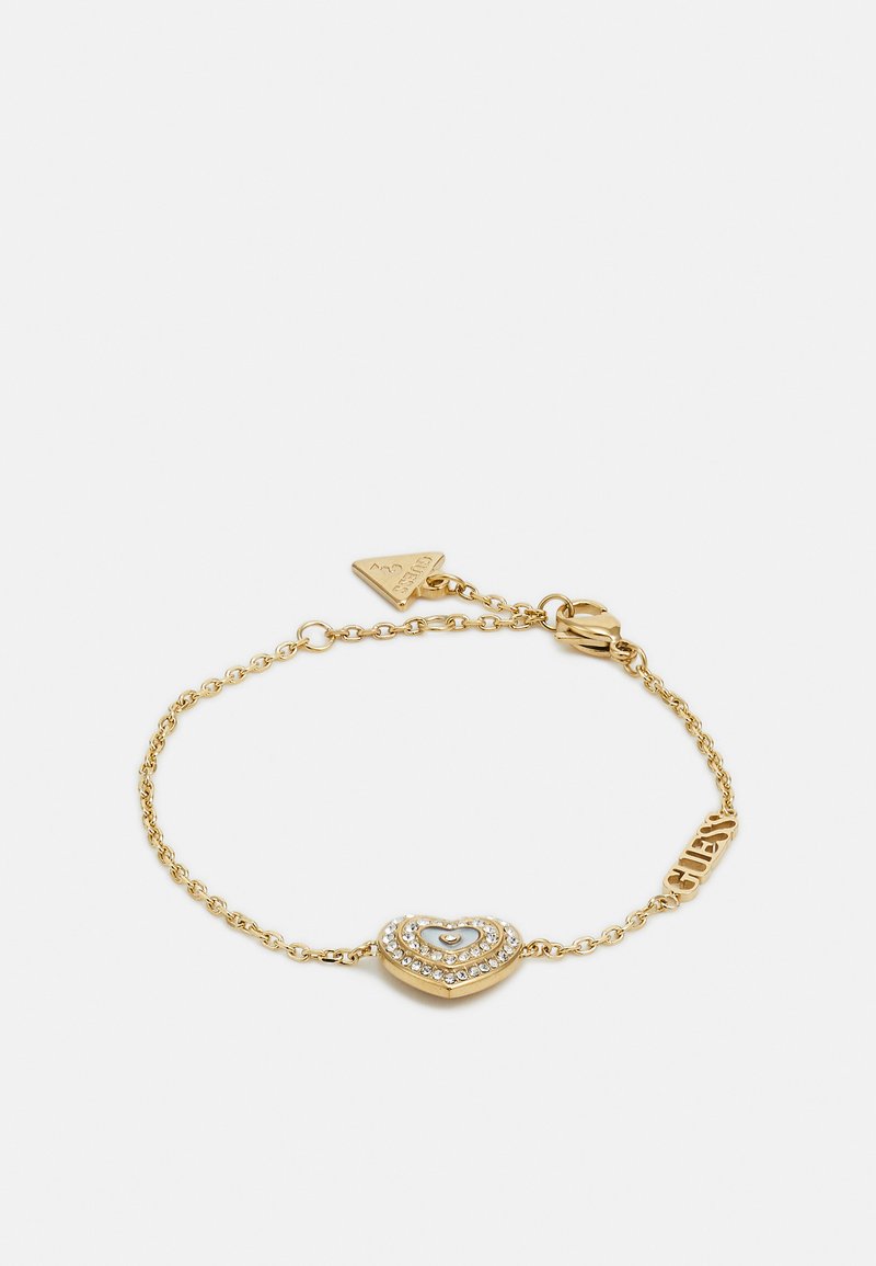 Gold-colored bracelet with a heart-shaped charm featuring layered design and embedded clear gemstones; marked with "Guess" on an accent.
