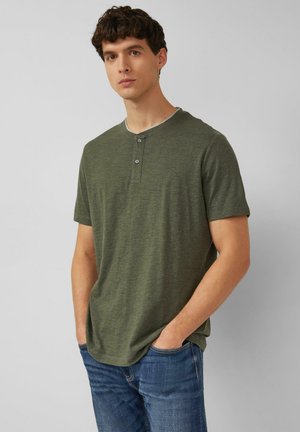 Young man wearing olive green short-sleeve henley shirt and blue jeans, standing with hands in pockets against plain gray background.