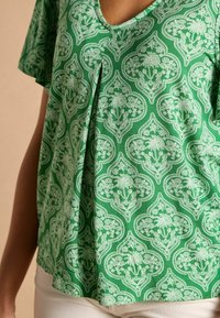 Zaļš rakstains tops ar V veida kakla izgriezumu, īsām piedurknēm un vieglu audumu ar baltiem ziedu motīviem un sarežģītu dizainu.