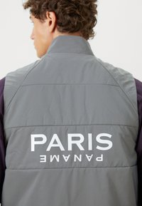 Gray padded vest featuring a high collar and prominent white text reading "PARIS PANAME" on the back. Smooth texture, modern design.