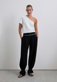 Weißes Oberteil mit einem Schulterträger, kombiniert mit lockeren schwarzen Hosen mit studs-verziertem Bund und schmal zulaufenden Knöcheln. Dazu werden minimalistische Sandalen getragen.