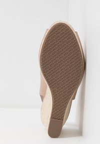 Vue de la semelle d'une sandale compensée beige avec semelle extérieure en caoutchouc texturé et talon plateforme recouvert de corde tressée, inclinée contre un mur blanc.