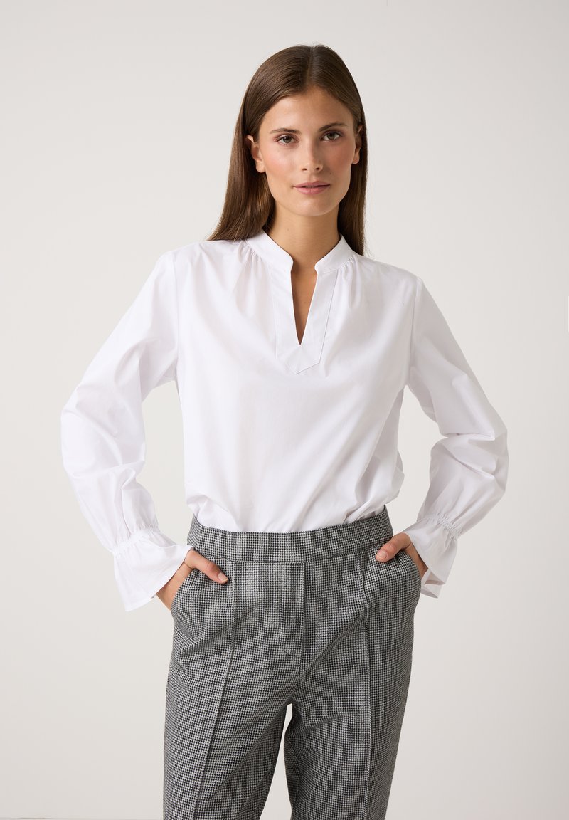 Weißer Langarm-Bluse mit V-Ausschnitt und gerafften Manschetten, kombiniert mit grauen karierten High-Waist-Hosen, die eine figurbetonte Silhouette zeigen.