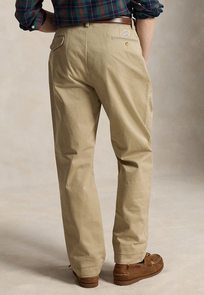 Man draagt beige chino's, een bruine leren riem, bruine mocassinschoenen en een groen-blauw geruit overhemd met opgerolde mouwen, gezien van achteren.