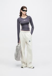 Γυναίκα με μαύρα γυαλιά ηλίου, γκρίζο μακρυμάνικο μπλουζάκι, λευκά παντελόνια φόρμας Nike, λευκά αθλητικά παπούτσια, holding γκρίζα τσάντα ώμου, στέκεται μπροστά σε λευκό φόντο.