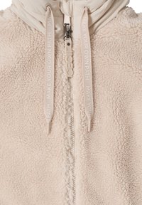 Fleece-takki vaaleassa beige-värissä, jossa on vetoketjusuljenta, teksturoitu kangas, joustavat nyörit painatuksella ja huppumalli.