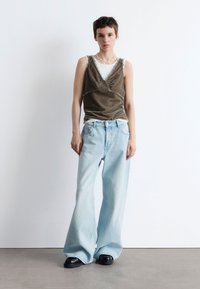 Braunes Samt-Top, das über einem weißen T-Shirt getragen wird, kombiniert mit hellblauen Schlagjeans und schwarzen Schuhen. Ein einfaches Collier als Akzent.