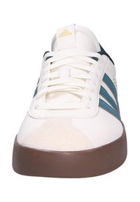 Zapatilla blanca con rayas verde azulado, parte superior de cuero texturizado y puntera de ante color crema. Presenta cordones blancos y una suela de goma marrón.