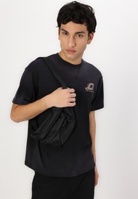 Black short-sleeve t-shirt with a round neckline and logo on the chest, paired with a textured black slouch bag held over the shoulder.
