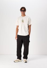 Camiseta de algodón crema con un gráfico verde y rosa, combinada con pantalones negros con herrajes plateados y un corte relajado. Zapatillas beige.