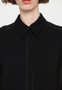 Blusa negra con un cuello estructurado, que presenta un panel frontal semitransparente y pliegues verticales. Textura suave y cierre de botones en el cuello.