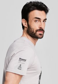 Jasnoszara koszulka z okrągłym dekoltem, z czarnymi nadrukami z przodu i na lewym rękawie. Rękaw posiada logo "Diverse™ ExtremeTeam" oraz grafikę Dakar.