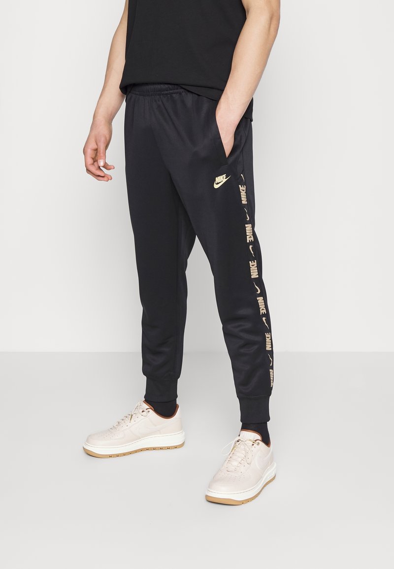 Hombre vistiendo pantalones deportivos negros de Nike con logotipos a lo largo del costado y zapatillas blancas, de pie contra un fondo claro y liso.