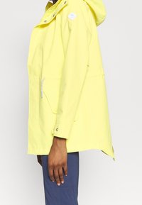 Gul regnjacka med huva, med en framficka, silverfärgade detaljer och en avslappnad passform. Mjuk, vattentät material.